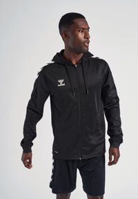 Felpa nera con zip realizzata in tessuto liscio, dotata di cappuccio, tasche frontali e logo/dettagli bianchi lungo le spalle. Abbinata a pantaloni corti neri.