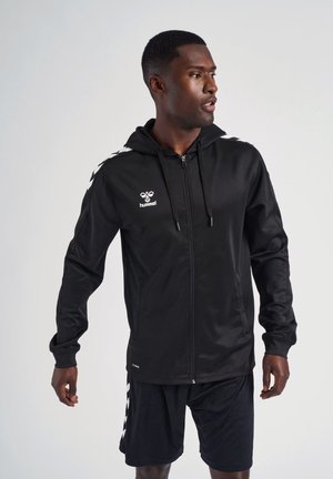 Schwarze Zip-Hoodie aus glattem Stoff, mit Kapuze, Fronttaschen und weißen Logos/Akzenten an den Schultern. Kombiniert mit schwarzen Shorts.
