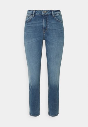 Siniset korkeavyötäröiset pillifarkut, joissa edessä nappi, vetoketju ja haalistuneet yksityiskohdat reisissä ja polvissa valkoista taustaa vasten.