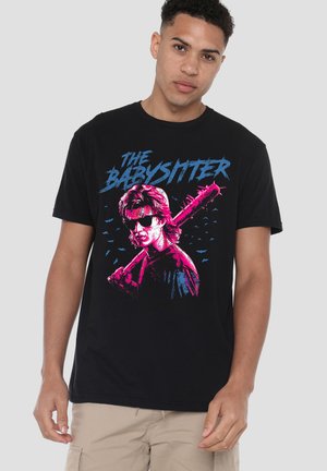Junger Mann trägt schwarzes T-Shirt mit neongrafik in Pink und Blau eines Menschen, der einen mit Stacheln besetzten Baseballschläger hält, und dem Text "The Babysitter."