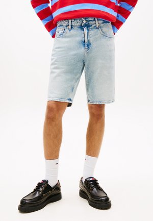 Person trägt hellblaue Jeans-Shorts, weiße knielange Socken, schwarze Lederschuhe und ein rot-blau gestreiftes Langarmshirt und steht.