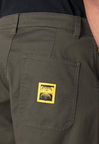 Olivgrüne Hose mit einem gelb-schwarzen Logo-Patch auf der Gesäßtasche. Der Stoff wirkt strapazierfähig, hat eine glatte Textur und ist im geraden Schnitt gestaltet.