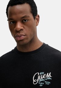 Camiseta negra de algodón con logotipo bordado de "Guess" en blanco y azul. Cuello redondo, mangas cortas, textura suave, diseño casual.