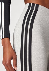 Grå träningstights med tre svarta vertikala ränder på sidorna. Har en åtsittande passform och ett mjukt, texturerat tyg.