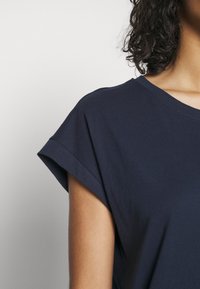 Personne portant une chemise bleu foncé à manches courtes, montrant son épaule et une partie de ses cheveux bouclés et foncés sur un fond clair uni.