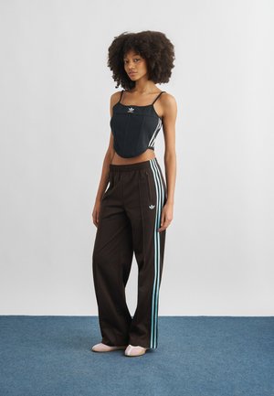 Mlada ženska z kodrastimi lasmi, oblečena v črno ohlapno zgornji del Adidas in črne Adidas trenirke s svetlo modrimi črtami, stoji na modrem preprogi.