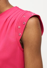 Ροζ αμάνικο τοπ με λεπτομέρειες από ασημί μεταλλικά κρικάκια κατά μήκος του ώμου, φορεμένο σε μπράτσο και ώμο με καφέ απόχρωση δέρματος.