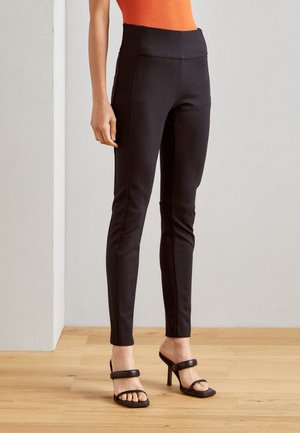 Zwarte high-waisted legging gemaakt van rekbare stof met stijlvolle zijnaad. Gecombineerd met zwarte haksandalen en een aansluitend oranje top.