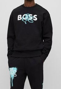 Sudadera negra con "BOSS" en letras blancas negrita y un diseño estilo graffiti en turquesa, con puños y dobladillo acanalados.