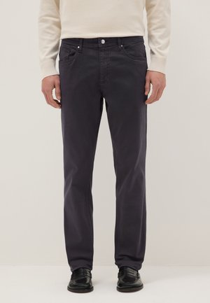 Man draagt zwarte jeans met rechte pijpen, zwarte loafers en een crèmekleurige trui met lange mouwen, staand tegen een egale lichte achtergrond.