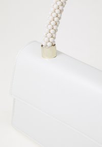 Biała skórzana torebka o usztywnionym kształcie. Posiada kształtowaną białą rączkę z koralików oraz złocony detal przy punkcie połączenia.