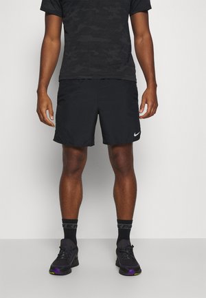 Homme portant un short de sport noir, une chemise noire à motifs, des chaussettes noires et des baskets noires, debout contre un mur uni.