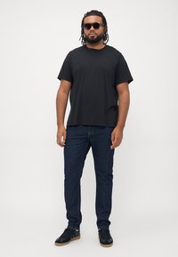 Svart kortermet T-skjorte kombinert med mørkeblå jeans og svarte sneakers. Modellen har på seg solbriller og står selvsikkert.