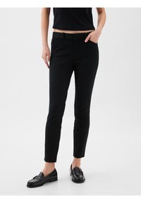 Personne portant un pantalon noir slim, des mocassins noirs et un haut noir court, debout avec une main dans la poche sur un fond blanc uni.