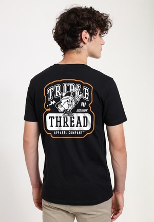 Młody mężczyzna o kręconych włosach, ubrany w czarną koszulkę z postacią kreskówkowego koszykarza i napisem "Triple Thread Apparel Company" na plecach.