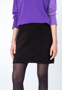Zwarte, gewatteerde minirok met ruitpatroon, gecombineerd met een paarse gebreide trui. Opaque zwarte maillots bedekken de benen.