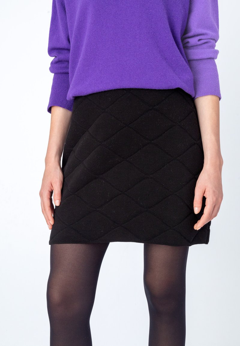 Zwarte, gewatteerde minirok met ruitpatroon, gecombineerd met een paarse gebreide trui. Opaque zwarte maillots bedekken de benen.