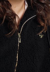 Svart fleecejacka med en texturerad yta, som har en guldglidare och ett litet märkesemblem. Närbild på kragen.