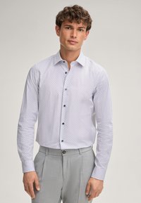Chemise blanche à boutons, ornée d'un motif géométrique en bleu marine, mise en valeur par des boutons foncés et des manches longues, associée à un pantalon gris clair.