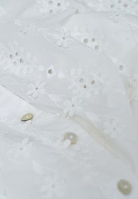 Tela blanca con bordado floral, detalles de ojales y botones tipo perla en una prenda ligera.