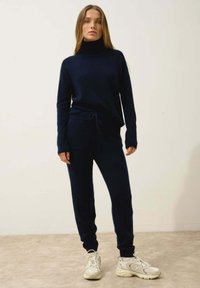 Mörkblå stickad loungeoutfit med höghalsskjorta och byxor med dragsko i midjan. Mjuk textur, figurnära design, matchat med ljusa sneakers.