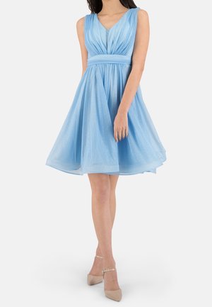 Robe bleu clair pailletée sans manches, longueur genou, avec un col en V et une taille cintrée, portée avec des talons beiges à bride à la cheville sur fond blanc.