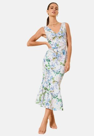 Frau mit Pferdeschwanz, die ein ärmelloses weißes Midikleid mit blauem und grünem Blumenmuster, eine goldene Halskette, Creolen und hautfarbene High Heels trägt.