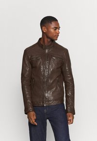 Giubbotto di pelle marrone con colletto alto, due tasche sul petto con zip e dettagli testurizzati. Indossato sopra jeans scuri, che mettono in risalto un design aderente.