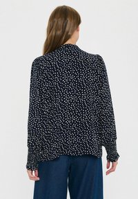 Tumesinine pluus valgete täppidega, pikkade varrukatega, nööpidega mansettide ja kaeluseta disainiga; lahtine lõike, sile tekstuur, minimalistlik stiil.