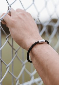 Musträpitud must käevõru metallist klambriga, kantud käel, mis haarab ketinfencesse. Lähedalt vaadatuna rõhutav tekstuur ja disain.