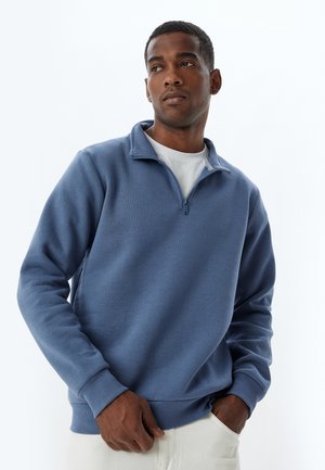Blauwe sweatshirt met rits van een textuur vergelijkbaar met fleece. Ribgebreide boorden en zoom, gedragen over een wit shirt. Eenvoudig, casual ontwerp.