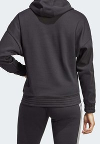 Sudadera con capucha negra, con puños y dobladillo acanalados. Fabricada en un tejido suave y texturizado. La vista trasera muestra el diseño sin logotipos ni gráficos visibles.