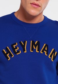 Sudadera azul con cuello redondo y texto bordado que dice "HEY MAN" en negro y amarillo con un acabado texturizado.