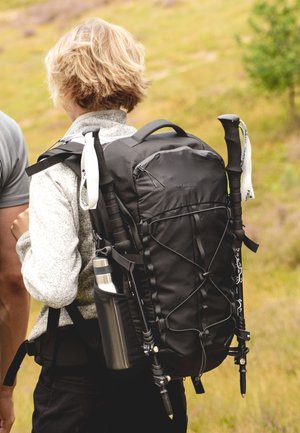 Black backpack with a textured exterior, adjustable straps, front pockets, and trekking poles attached; features a side pocket for a silver water bottle.