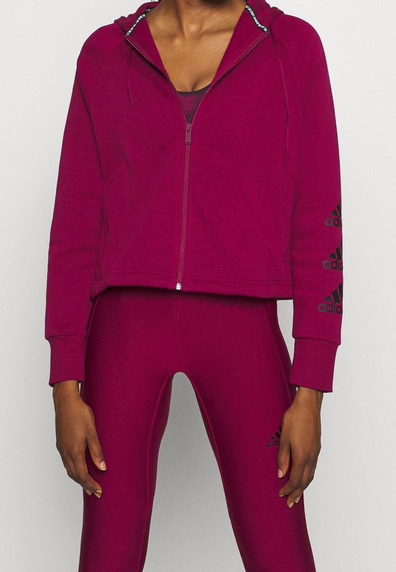 Femme portant un sweat à capuche zippé rose foncé assorti et des leggings avec des logos Adidas noirs sur les manches et la cuisse, debout sur un fond blanc.
