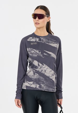 Kvinde iført mørkegrå og hvide mønstrede cykeltrøje med lange ærmer, sorte cykelshorts og store wraparound-solbriller.