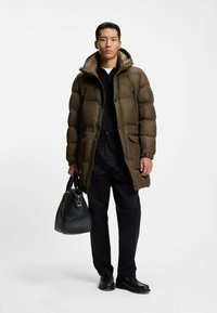 Olivgrüner Puffermantel mit Kapuze, mit Reißverschluss vorne und seitlichen Taschen, getragen über einem schwarzen Shirt und einer Hose. Schwarze, strukturierte Tasche in der Hand.