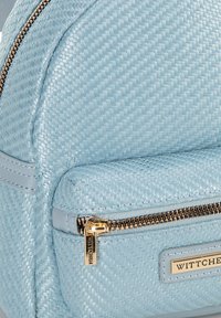 Mochila de color azul claro hecha de material tejido con un acento de cuero suave, que presenta un zipper dorado y una etiqueta de marca. Diseño redondeado.