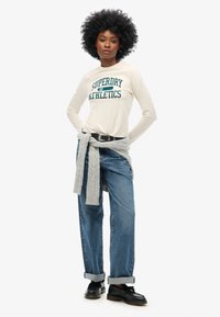 Kremowa koszula z długim rękawem z napisem "Superdry Athletics" w kolorze zielonym, zestawiona z jasnoniebieskimi dżinsami o prostym kroju i szarym swetrem przewiązanym w talii.