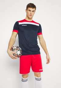 Marineblaues Trikot mit roten Ärmeln und weißem Streifen, kombiniert mit roten Shorts. Der Spieler hält einen schwarz-weißen Fußball und trägt weiße Socken mit Streifen.