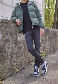 Chaqueta acolchada verde con capucha sobre una camiseta negra, combinada con jeans oscuros y zapatillas blanco y negro, de pie contra una pared de hormigón.