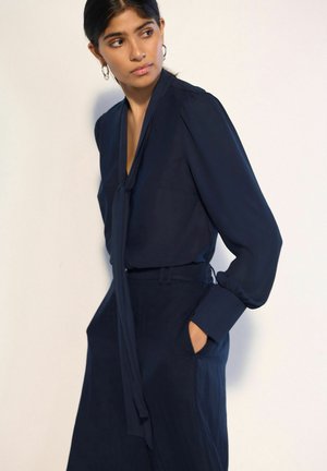 Donna che indossa una camicetta blu scuro a maniche lunghe con collo a nodo e pantaloni coordinati a vita alta, in piedi con una mano in tasca.