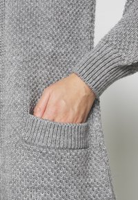 Szary dzianinowy kardigan z fakturowanym wzorem, ściągaczowymi mankietami i przednią kieszenią. Zbliżenie na dłoń spoczywającą w kieszeni.