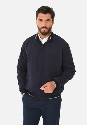 Uomo con barba che indossa una giacca blu scuro con cerniera sopra una maglietta bianca e pantaloni blu scuro, in piedi davanti a uno sfondo chiaro e semplice.