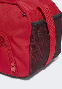Bolsa de tela roja con una superficie texturizada, presenta un lado de malla negra, cierre de cremallera y un asa roja resistente. Diseño compacto y práctico.