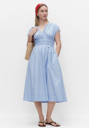 Kvinde iført en midi kjole med blå og hvide striber, rød hovedbeklædning, bærende en flettet taske og iført sorte sandaler, stående mod en hvid baggrund.