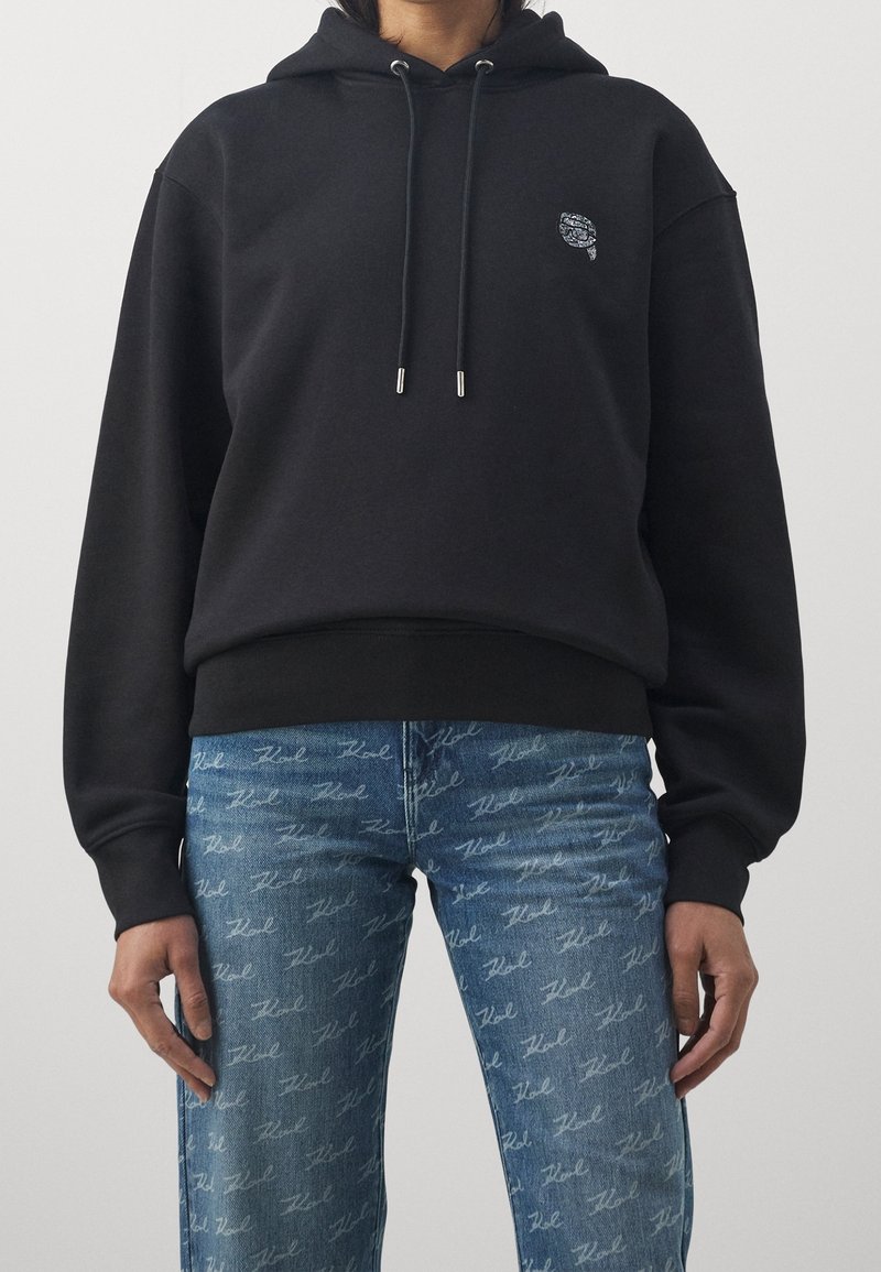 Personne portant un sweat à capuche noir avec cordons et un jean bleu avec un motif intégral en écriture cursive blanche.