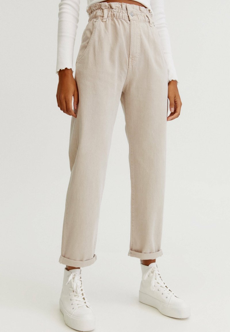 Pantalones de mezclilla beige con cintura elástica, bolsillos laterales y puños enrollados, combinados con zapatillas altas blancas.