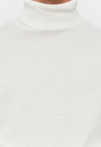 Primer plano de una persona que lleva un suéter de cuello alto de punto blanco liso, mostrando el cuello y la parte superior del pecho.