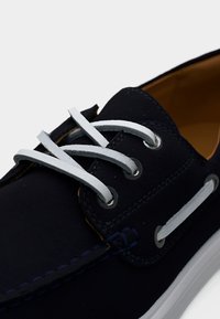 Σκούρο μπλε ναυτικό παπούτσι με λευκά δερμάτινα κορδόνια περασμένα μέσα από μεταλλικά οιλέτα και λευκή λαστιχένια σόλα, σε κοντινή λήψη.